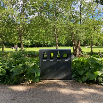 En elegant, cylinderformet affaldsspand fra Peoples i mørkt stål, placeret på et flisebelagt torv foran en moderne bygning.