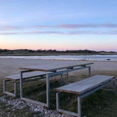 En robust bænk fra Peoples med stålramme og træsæde, placeret i en park med grønne træer i baggrunden.