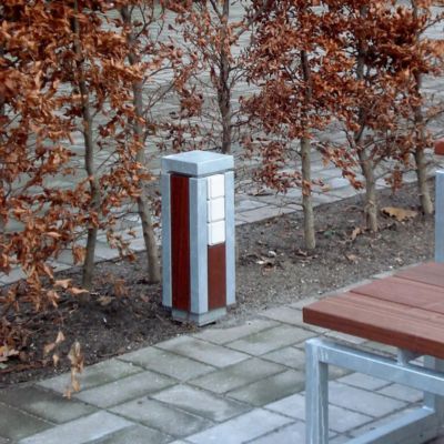 En stilren el-pullert i børstet stål fra Peoples, diskret placeret ved siden af en bænk på et torv.